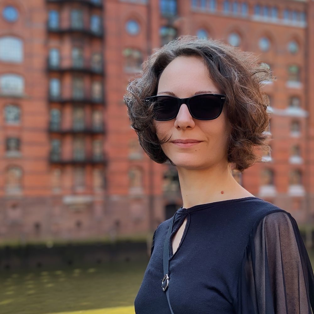 Frau mit braunen Locken und Sonnenbrille steht vor einem Haus in der Hamburger Speicherstadt.