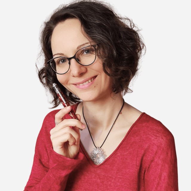 Frau mit braunen Locke, Brille und einem Kuli in der Hand lächelt.