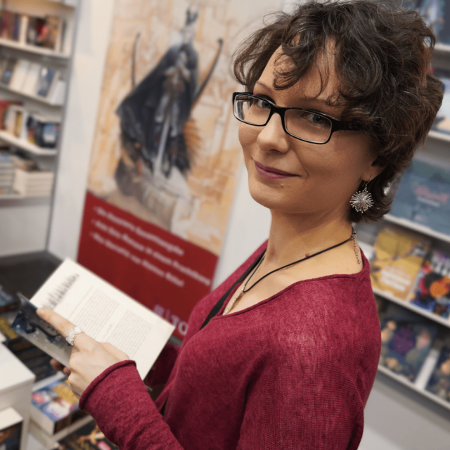 Frau mit braunen Locke, Brille und einem Buch in der Hand lächelt.