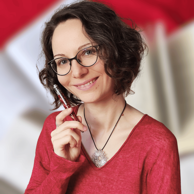 Frau mit braunen Locke, Brille und einem Kuli in der Hand lächelt.